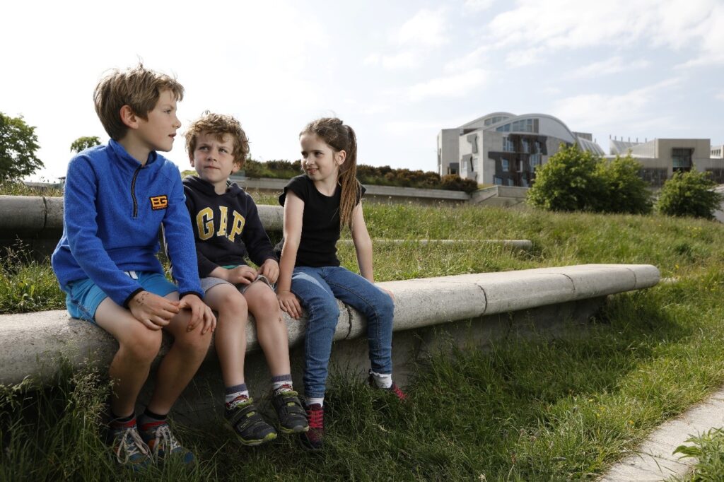 Three children sitting on a wall with the Scottish Parliament in the background